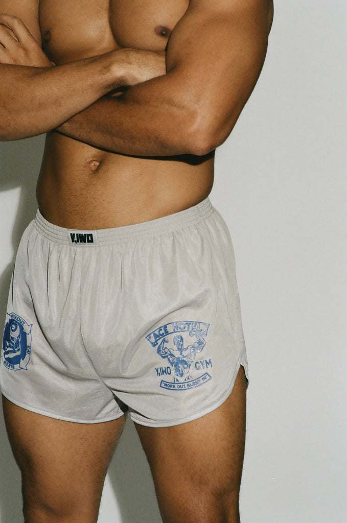 Close-up of a shirtless muscular male model wearing Y,IWO x Ace Hotel grey microfiber quad shorts with blue bodybuilding graphics, short inseam, and curved side hems that expose the thighs.