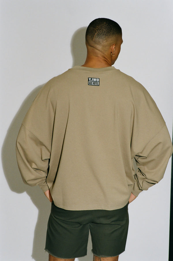 Back view of a male model wearing the Y,IWO x Ace Hotel oversized long sleeve T-shirt in stone beige with dark athletic shorts, showing the relaxed fit, dropped shoulders, and small collaboration logo below the neckline.